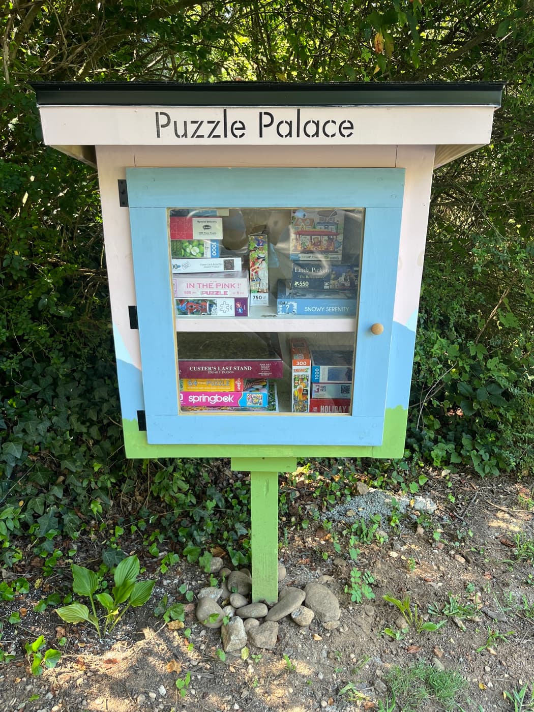 The Puzzle Palace: a pink box with a dark roof, a stencil-cut Puzzle Palace sign, and a periwinkle glass door over two shelves of puzzle boxes.
